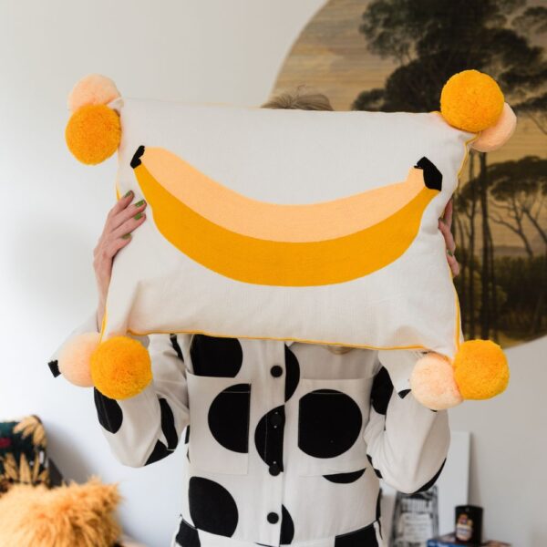 person holding up a white cushion with large embroidered yellow banana on with yellow large pom poms pompom