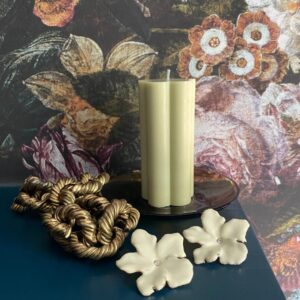 light sage green flower shaped soy wax pillar candle on a mantlepiece with jewellery and floral wallpaper background