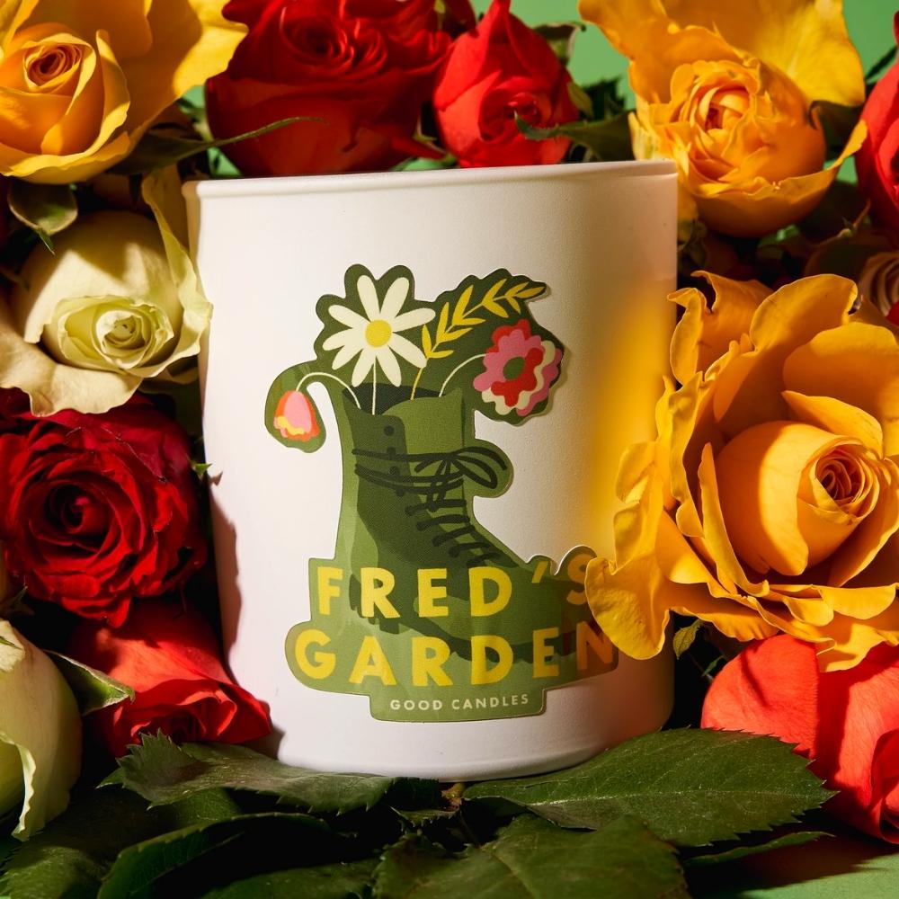 Soy wax candle with boots and flowers and text that says Fred's Garden on a background of roses