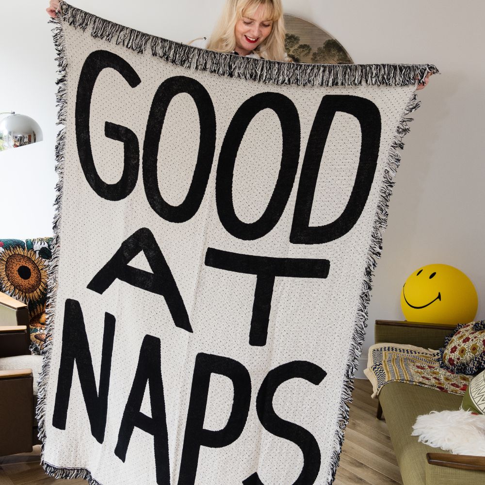 Living room setting with person holding up a monochrome fringed throw blanket that says good at naps on in large text
