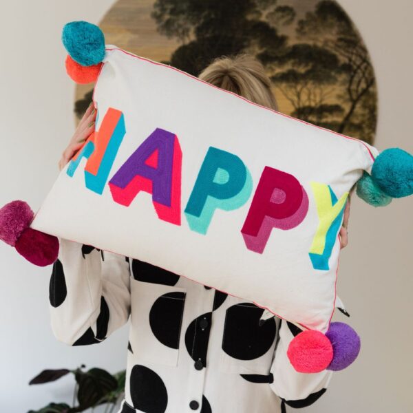 Person holding up a ivory cushion made from PET recycled bottles that has the word Happy emvroidered on the front in multicoloured letters and large pom poms
