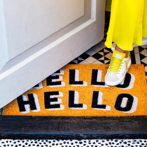 a foot wearing white and yellow trainers standing on a sustainable eco door mat with black and white text saying hello hello