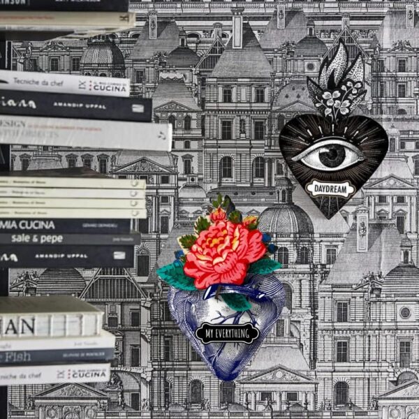 image of wall with printed building wall paper with a stack of books and two exvoto decorative wall hanging mexican style hearts one is an anatomical heart with a rose and says my everything and the other has the all seeing eye and says daydream