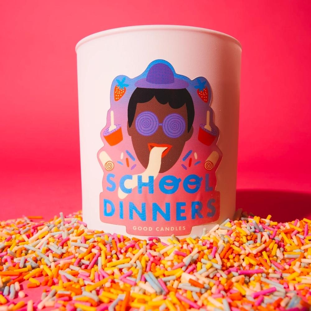 school dinners sweets scented white candle with a face with a hat and glasses on with tongue out and words, on a pink background with hundred and thousands candy