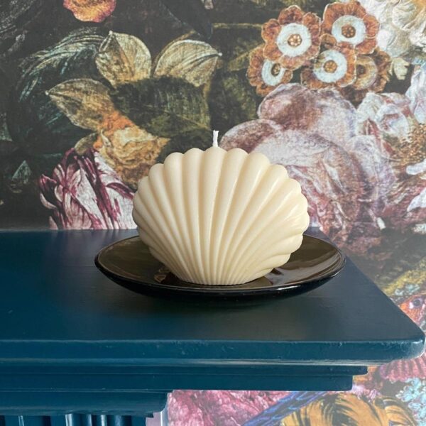 ivory cream sea shell shaped candle on a glass dish on a mantlepiece with floral wallpaper background