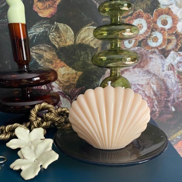 natural nude sea shell shaped candle on a glass dish on a mantlepiece with jewellery and floral wallpaper background