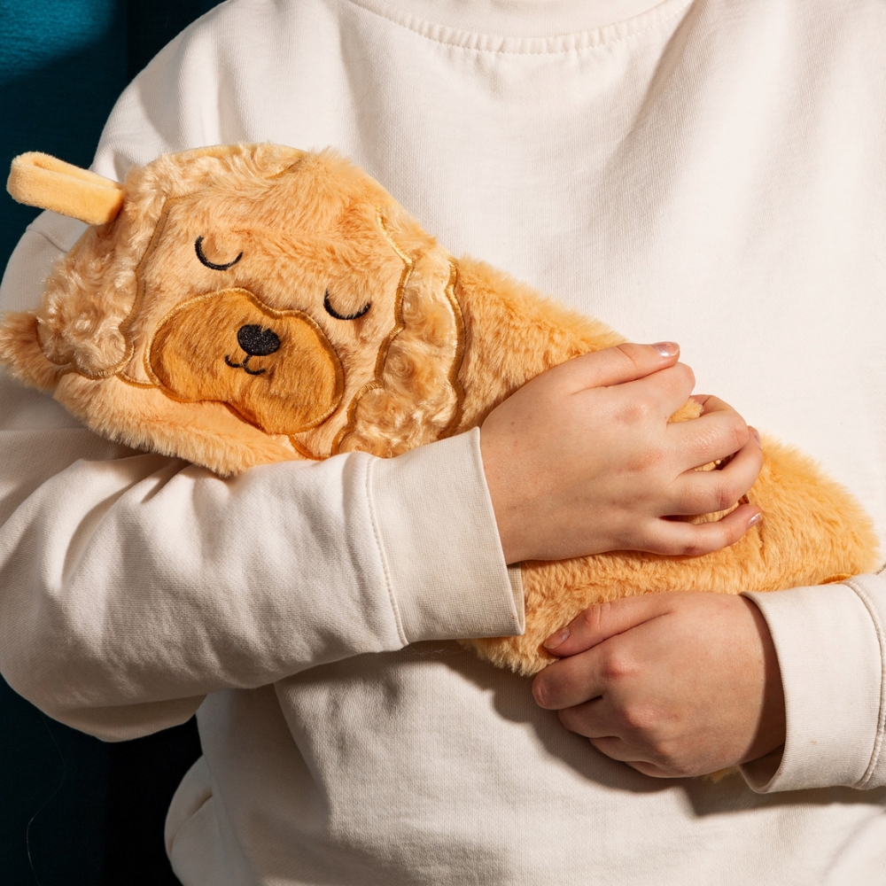 A PERSON HOLDING A FAUX FUR HUGGABLE ANIMAL SHAPED HOT WATER BOTTLE IN THE SHAPE OF A GOLDEN COCKAPOO DOG