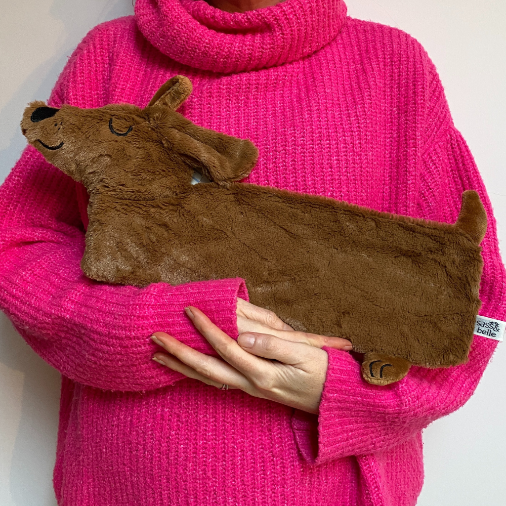 A PERSON HOLDING A FAUX FUR HUGGABLE DOG SHAPED HOT WATER BOTTLE IN THE SHAPE OF A BROWN DACHSHUND SAUSAGE DOG