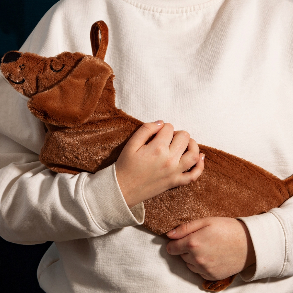 A PERSON HOLDING A FAUX FUR HUGGABLE DOG SHAPED HOT WATER BOTTLE IN THE SHAPE OF A BROWN DACHSHUND SAUSAGE DOG