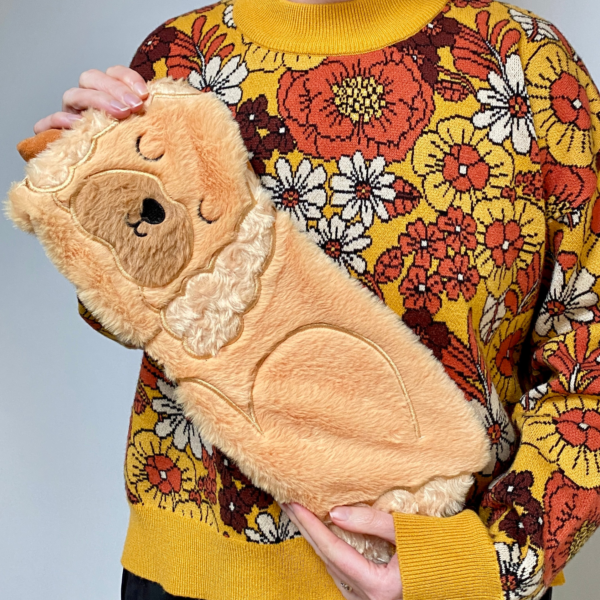 A PERSON HOLDING A FAUX FUR HUGGABLE ANIMAL SHAPED HOT WATER BOTTLE IN THE SHAPE OF A GOLDEN COCKAPOO DOG