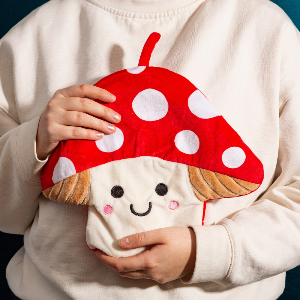 A PERSON HOLDING A HUGGABLE CUTE RED AND WHITE MUSHROOM WITH SMILEY FACE TOADSTOOL SHAPED HOT WATER BOTTLE